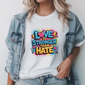 Può includere: T-shirt bianco con la scritta "LOVE IS STRONGER THAN HATE" in un design colorato in stile graffiti. Il design include un cuore rosa e fulmini, con uno sfondo di schizzi di vernice.