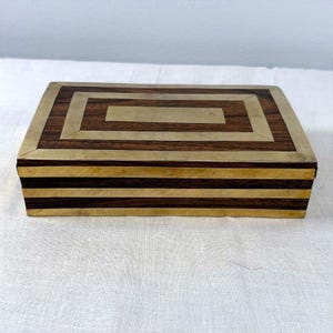 May include: A rectangular wooden box featuring dark brown wood grain panels accented with polished brass inlay. The lid displays a geometric brass border and a central rectangular brass plate, while the sides feature horizontal brass stripes.