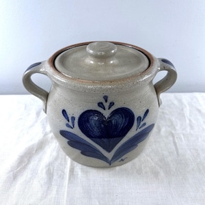 May include: A speckled beige stoneware crock with two side handles and a matching lid. The front features a hand-painted dark blue heart motif with stylized leaf accents. The rim and handle edges are accented with a thin brown glaze line.