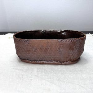 May include: An oval-shaped ceramic planter with a dark brown, glossy glaze. The exterior surface features a repeating, embossed honeycomb or circular dot pattern. The rim is smooth and rounded, while the base has a slightly irregular, textured edge.