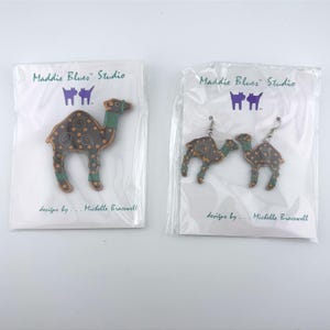 May include: A brown camel-shaped brooch and a pair of matching camel-shaped earrings, both featuring green accents and yellow polka dots. Each item is packaged in a clear plastic sleeve with a white card labeled Maddie Blues Studio.
