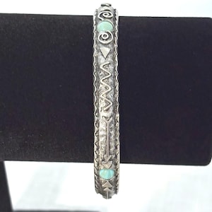 May include: A silver-toned metal bangle bracelet featuring a textured, oxidized finish. The surface is decorated with engraved geometric patterns, including zigzags and arrows, interspersed with two small, smooth, light blue circular cabochons.