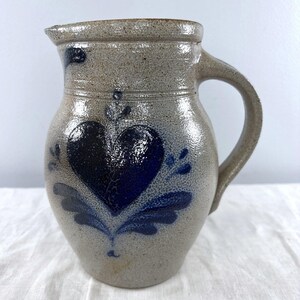 May include: A salt-glazed stoneware pitcher with a speckled beige finish and a prominent cobalt blue heart motif painted on the front. The vessel features a rounded body, a sturdy side handle, and a small pouring spout at the top rim.