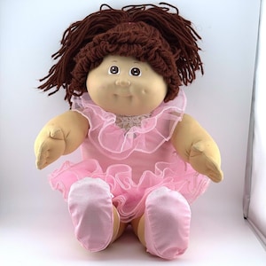 May include: A soft-sculpture doll with dark brown yarn hair, a round face, and dimpled cheeks. The doll wears a light pink sleeveless outfit featuring a ruffled tulle collar and a matching ruffled skirt, paired with soft pink fabric shoes.