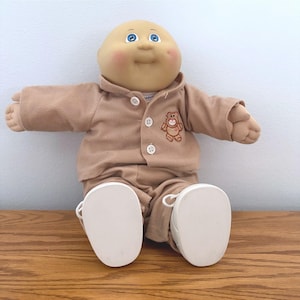 May include: A doll with a round, tan-colored head, blue eyes, and rosy cheeks. The doll wears a tan long-sleeved collared shirt with three white buttons and a small brown teddy bear embroidery on the chest, paired with matching pants and white shoes.