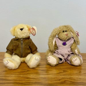 May include: A cream-colored plush bear wearing a brown zippered jacket and a tan plush rabbit wearing lavender corduroy overalls. Both stuffed animals feature a heart-shaped Ty tag attached to their ears, with the bear on the left and the rabbit on the right.