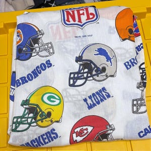 May include: A white fabric sheet featuring a repeating pattern of various NFL football team helmets and logos, including the Broncos, Lions, Packers, and Chiefs. The print includes the NFL shield logo and the text TM 1995 NFLP in black ink.