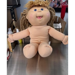 May include: A vintage Cabbage Patch Kids doll with a peach-colored body, arms, and legs. The doll has a round face with blue eyes, rosy cheeks, and a smiling mouth. Its light brown hair is styled in a curly, frizzy fashion. The doll is sitting on a reflective surface.
