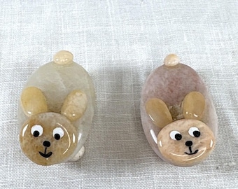 May include: Two oval-shaped stone figurines shaped like rabbits. Each figurine features a smooth, polished body, two small ears, and a painted face with white eyes, black pupils, and a black nose and mouth. The stones are light beige and tan.