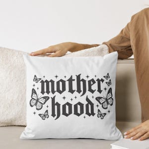May include: White square pillow with the word "motherhood" in a gothic font, surrounded by butterfly illustrations and star accents in black. The pillow is on a neutral-toned surface.