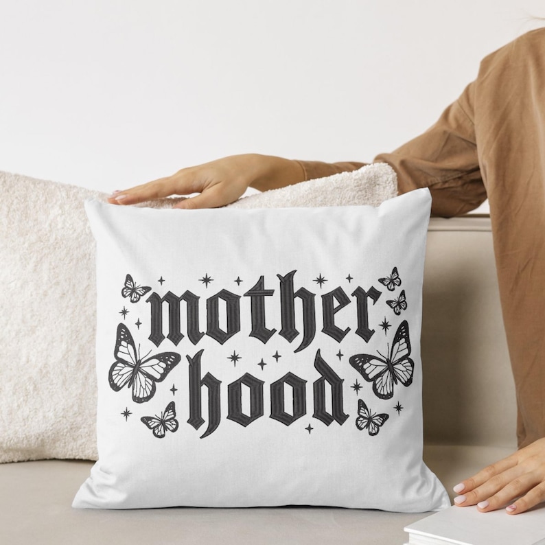 May include: White square pillow with the word "motherhood" in a gothic font, surrounded by butterfly illustrations and star accents in black. The pillow is on a neutral-toned surface.