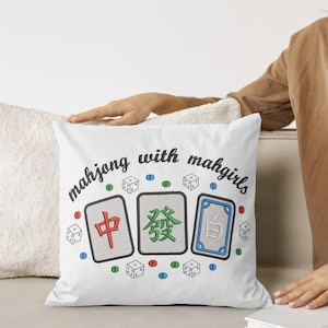 May include: White square pillow with embroidered Mahjong tiles and dice. The pillow has the text "mahjong with mahgirls" in a script font. The Mahjong tiles are red, green, and blue with black borders. Scattered around the tiles are small dice and colored dots.