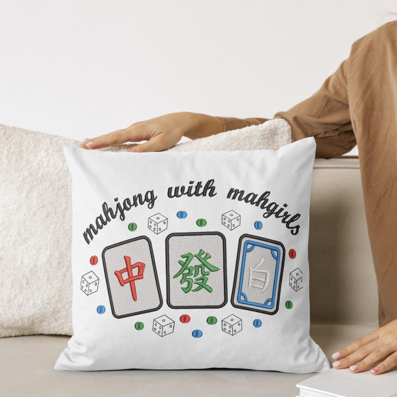 May include: White square pillow with embroidered Mahjong tiles and dice. The pillow has the text "mahjong with mahgirls" in a script font. The Mahjong tiles are red, green, and blue with black borders. Scattered around the tiles are small dice and colored dots.