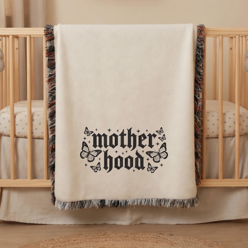 May include: Cream-colored baby blanket with the word "motherhood" in a gothic font, surrounded by butterfly illustrations. The blanket has a fringed edge with brown, black, and orange threads. The blanket is draped over a wooden crib.
