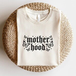 May include: Cream-colored t-shirt with the word "motherhood" in a gothic font, adorned with butterfly illustrations. The shirt is made of soft material and is displayed on a woven surface. The design includes stars and butterflies.