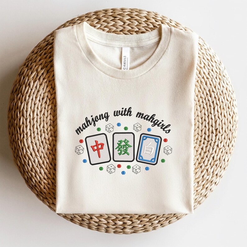 May include: Cream-colored t-shirt featuring the text "mahjong with mahgirls" above three mahjong tiles and dice. The tiles are red, green, and blue, with Chinese characters. The shirt is displayed on a woven surface.