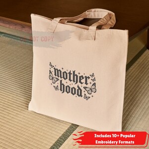 May include: Beige canvas tote bag with the word "motherhood" in a gothic font, decorated with butterfly illustrations. The bag has two handles and is sitting on a woven mat. The bottom right corner has a red banner with white text.
