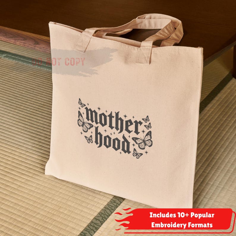 May include: Beige canvas tote bag with the word "motherhood" in a gothic font, decorated with butterfly illustrations. The bag has two handles and is sitting on a woven mat. The bottom right corner has a red banner with white text.