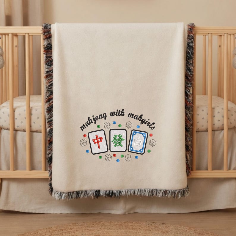 May include: Cream-colored baby blanket with a fringed edge, draped over a wooden crib. The blanket features embroidered Mahjong tiles and the text "mahjong with mahgirls" in a playful font. The design includes dice.