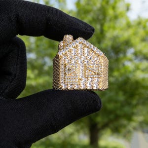 May include: A gold-colored house-shaped ring encrusted with numerous clear, sparkling gemstones. The ring features a detailed design, including a roof, windows, and a chimney. The ring is held against a blurred green background.