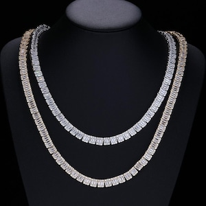 May include: Two necklaces on a black display stand. One necklace is gold-toned, the other is silver-toned. Both necklaces feature a series of rectangular, clear stones. The necklaces are displayed against a black background.
