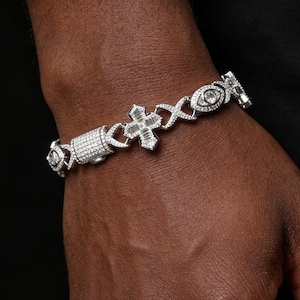 May include: A silver bracelet with a cross and eye design. The bracelet is encrusted with small, clear gemstones. The cross is the focal point, with a baguette-cut gemstone design. The bracelet is on a dark background.