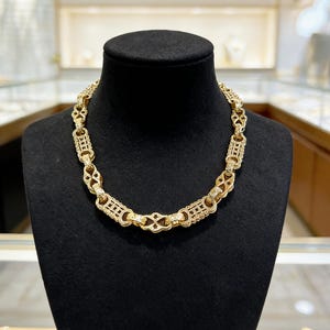 May include: A gold-colored necklace with an intricate, geometric design. The necklace is displayed on a black mannequin. The chain links feature a repeating pattern of rectangular and diamond-shaped elements, creating a luxurious look.