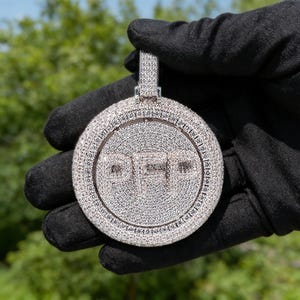 May include: A round, silver-colored pendant with the letters "PEP" encrusted with small, sparkling stones. The pendant has a loop at the top for a chain and is held against a blurred green background.