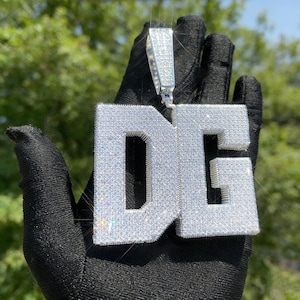 Puede incluir: Colgante plateado con las letras "DG" incrustadas con pequeñas piedras brillantes. El colgante está suspendido de una fianza plateada, sostenido por una mano enguantada sobre un fondo verde borroso.