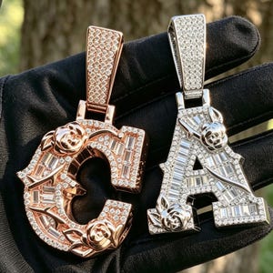 Pode incluir: Dois pingentes em forma de letras C e A, um em ouro rosa e outro em tom prateado. Ambos apresentam pedras incrustadas de corte baguette e redondo, com motivos esculpidos de rosas e espinhos integrados na superfície do metal.
