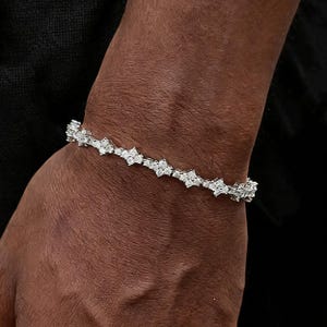 Pode incluir: Uma pulseira prateada com um design floral. A pulseira é feita de pequenos elementos em forma de estrela ligados entre si. A pulseira é usada no pulso, contra um fundo escuro.