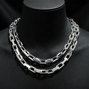 May include: A double-layered silver-toned chain necklace featuring rectangular, elongated links. The polished metal links are interconnected to form a uniform, structured chain design that rests around the neck, showcasing a bright, reflective finish.