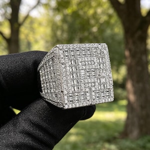 May include: A large, square-shaped silver-tone ring featuring a prominent letter R on the face. The entire surface of the ring is densely encrusted with numerous small, clear, rectangular baguette-cut and round-cut stones that create a reflective finish.