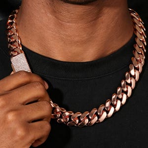 May include: A thick rose gold-toned cuban link chain necklace featuring a rectangular clasp encrusted with numerous small, clear, reflective stones. The chain consists of interlocking, polished, rounded metal links that sit around the neck.