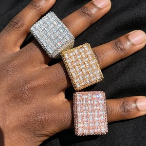 May include: Three rectangular rings featuring a grid of baguette-cut clear stones. One ring is silver-toned, one is gold-toned, and one is rose gold-toned. Each ring has a wide, flat top surface completely covered in small, sparkling, rectangular stones.