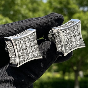 May include: A pair of square-shaped silver cufflinks. Each cufflink is encrusted with numerous small, clear gemstones, creating a sparkling effect. The cufflinks have a polished, reflective surface, and are set against a blurred green background.