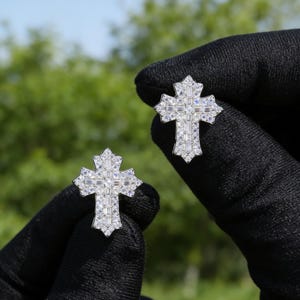 May include: A pair of silver-toned cross-shaped earrings. Each earring is encrusted with multiple small, clear, rectangular-cut stones. The earrings are held against a blurred green background.