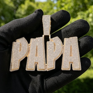 May include: A gold-colored pendant spelling out the word "PAPA" in capital letters. The letters are encrusted with small, sparkling stones. The pendant is held against a blurred green background.
