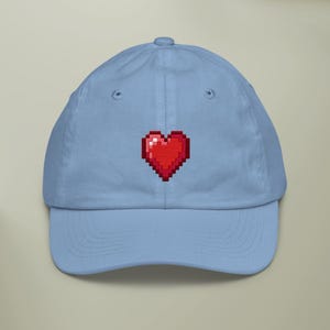 May include: A light blue baseball cap featuring a pixelated red heart design. The cap has a curved brim and an adjustable strap at the back. The heart design is centered on the front of the cap.