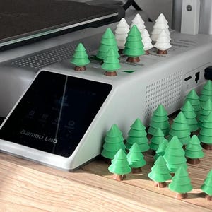 May include: A Bambu Lab 3D printer base unit featuring a black touchscreen display with the brand name printed in white. The top and surrounding area are decorated with numerous small, three-dimensional green and white plastic pine trees.