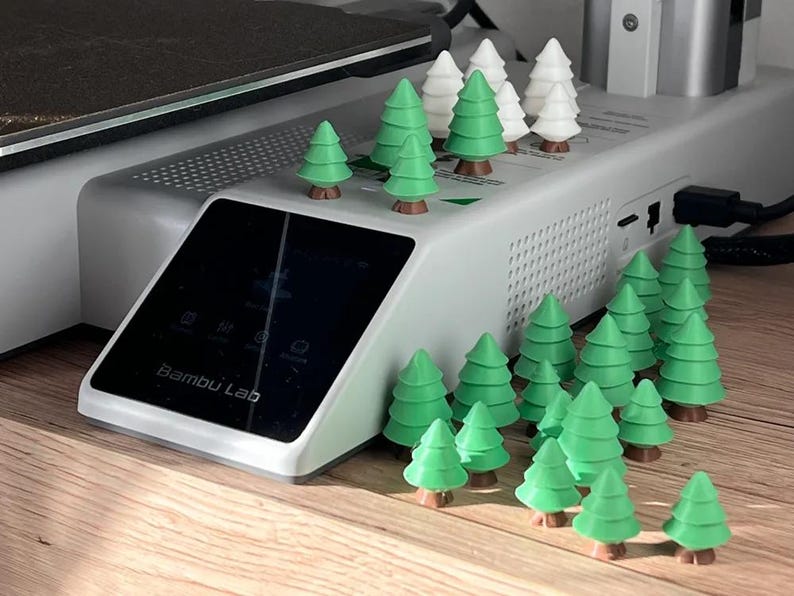 May include: A Bambu Lab 3D printer base unit featuring a black touchscreen display with the brand name printed in white. The top and surrounding area are decorated with numerous small, three-dimensional green and white plastic pine trees.