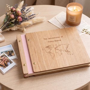 May include: A wooden photo album with a light wood grain cover featuring an engraved world map and the text "The Adventures of Felix+Maeve". The album has a light pink spine secured with gold-colored metal rivets and contains internal pages.