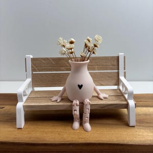 Könnte beinhalten: Eine skurrile rosa Vase mit Armen und Beinen, einem schwarzen Herz und einem Strauß getrockneter weißer Blumen sitzt auf einer Miniatur-Holzbank mit weißen Akzenten. Die Bank steht auf einer Holzoberfläche.