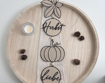 May include: A vertical hanging decoration featuring a wooden leaf, a circle with the text Herbst, a pumpkin, and a circle with the text liebe, all connected by wooden beads on a white lace ribbon, accompanied by small pinecones and a tea light.
