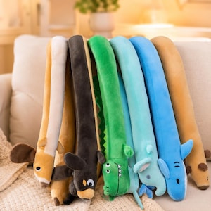 Op de afbeelding: Een verzameling lange, cilindervormige pluche dieren naast elkaar. De rij bevat een beige en witte hond, een donkerbruine hond, een groene krokodil met tanden, een lichtblauwe olifant, een felblauwe haai en een beige dierfiguur.