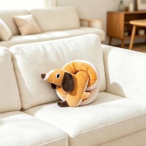 Op de afbeelding: Een knuffel in de vorm van een opgerolde teckel met een beige lichaam, donkerbruine oren en een witte snuit. Het zachte pluche dier heeft een rond, gedraaid ontwerp met een gladde textuur en een gestileerde honden kop met zwarte ogen.