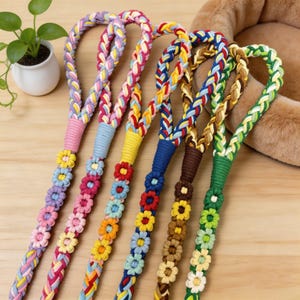 Op de afbeelding: Een verzameling kleurrijke gevlochten hondenriemen. Elke riem heeft een ander kleurenschema, waaronder roze, blauw, geel en groen, met een bijpassend bloemendesign. De riemen zijn gemaakt van een duurzaam, geweven materiaal.