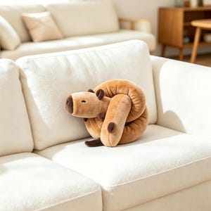 Op de afbeelding: Een beige pluche capibara-speeltje in de vorm van een opgerolde knoop. Het knuffeldier heeft een donkerbruine neus, kleine donkerbruine oren en donkerbruine voetjes. De zachte, gestructureerde stof is lichtbruin met een gladde, ronde uitstraling.