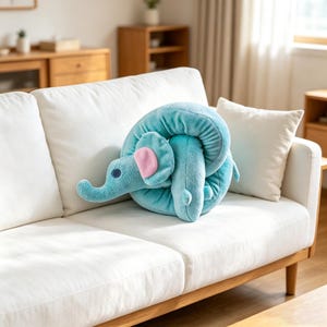 Op de afbeelding: Een pluche knuffeldier in de vorm van een opgerolde olifant. Het speelgoed is gemaakt van zachte, lichtblauwe stof met een gladde textuur. Het heeft een slurf, een klein roze binnenoor en een donkerblauw cirkelvormig oogdetail op de zijkant.