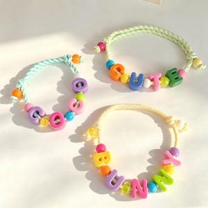 Könnte beinhalten: Drei Armbänder aus geflochtenen hellgrünen und gelben Kordeln mit bunten Kunststoff-Buchstabenperlen und kleinen runden Akzentperlen. Die Armbänder zeigen die Wörter COCO, CUTE und BUNNY in verschiedenen leuchtenden Farben wie Pink, Orange und Blau.